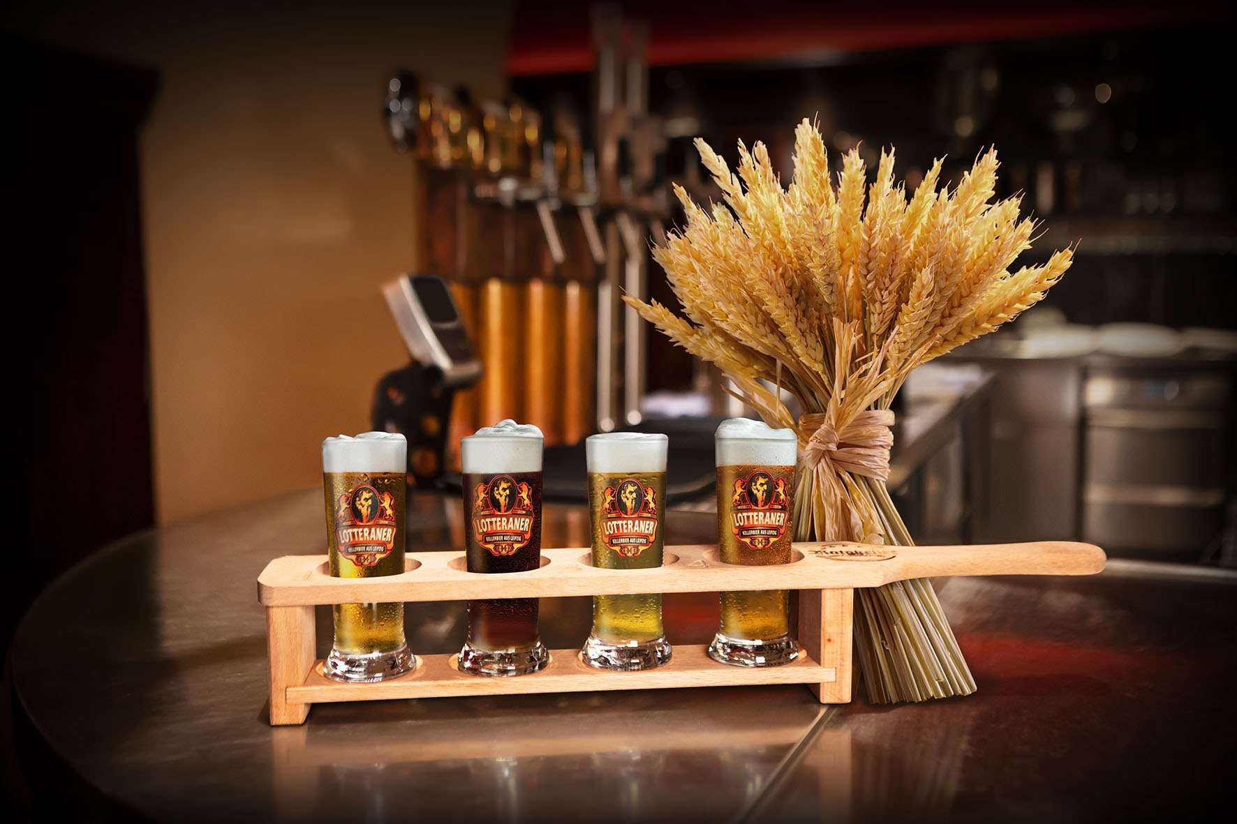 Vier Gläser Lotteraner Bier in Holzträger neben einem Bündel Getreide auf einer Theke.